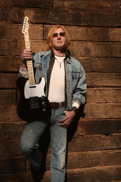 Clayton Mack, country rock musician, holding an electric guitar in a denim jacket against a wooden wall